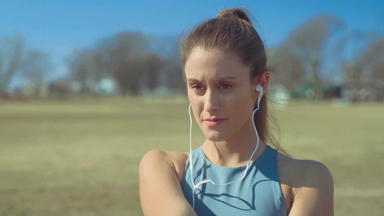 retrato de una joven poniéndose auriculares para escuchar música, luego comienza a estirarse para calentarse para su entrenamiento al aire libre en un día soleado