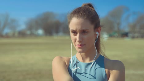 portrait of young woman putting on earphones to listen to music, then begins stretching to warm up for her outdoor workout on sunny day