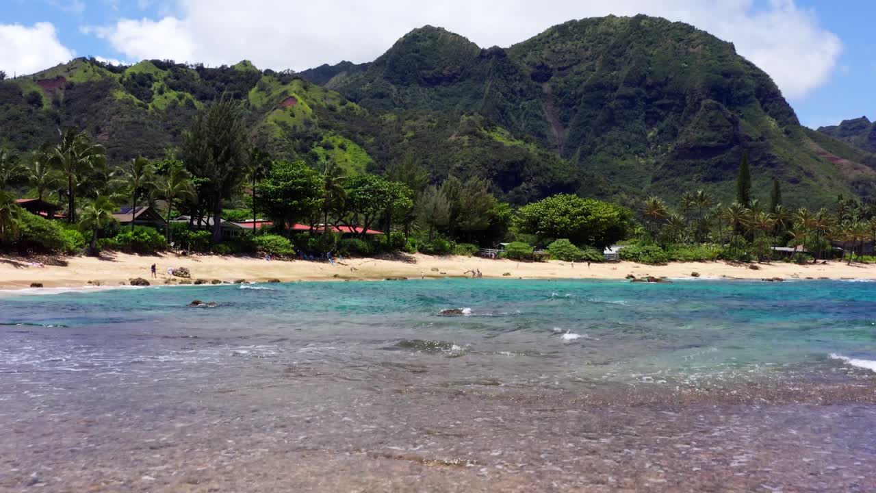 Drone shot from above Tunnels Beach on Kauai’s North Shore. Bright coral reef system and snorkelers are visible below, with green mountains rising in the background. Clear, sunny day