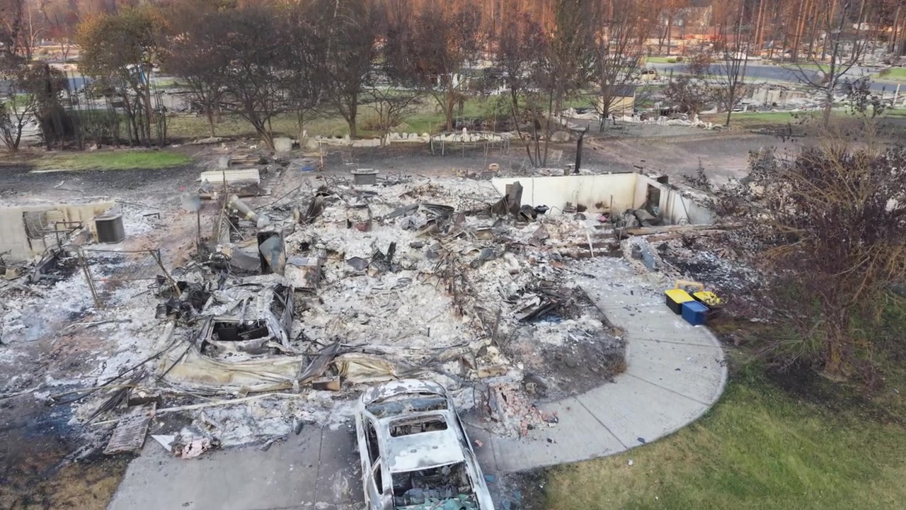 Danni dell'incendio di Spokane che mostrano una ripresa aerea bassa all'indietro su lotti bruciati, con terreno carbonizzato, detriti e distruzione diffusa dopo l'incendio del 2026