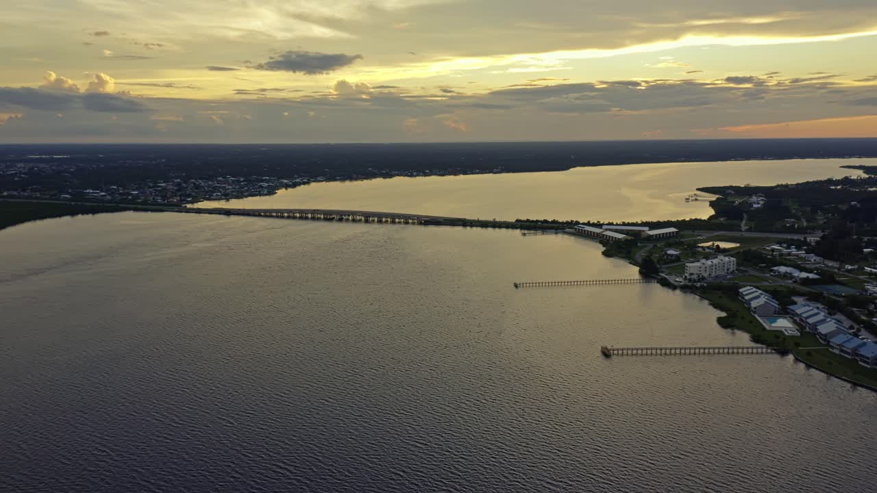 A bridge crosses expansive bay waters as the horizon glows with the warm hues of sunset, casting reflections across the calm surface and outlining the distant shoreline