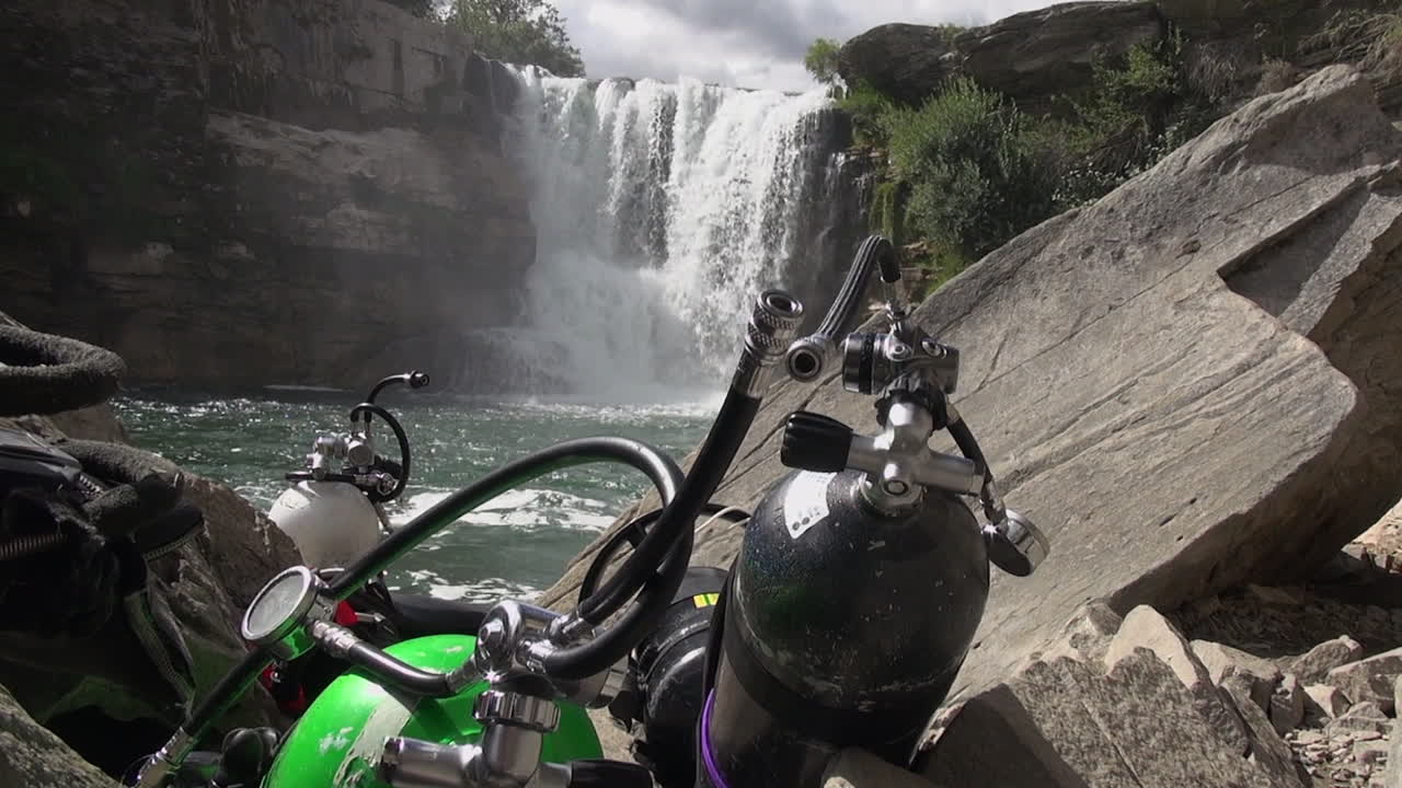 Bombole subacquee, attrezzatura su rocce a bordo fiume sotto una cascata fluviale turbolenta.