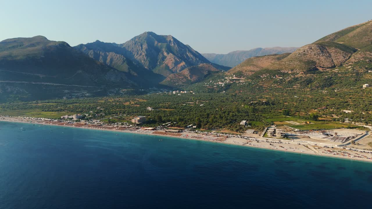 A beautiful coastline with clear blue water, beachgoers, and towering mountains, aerial view