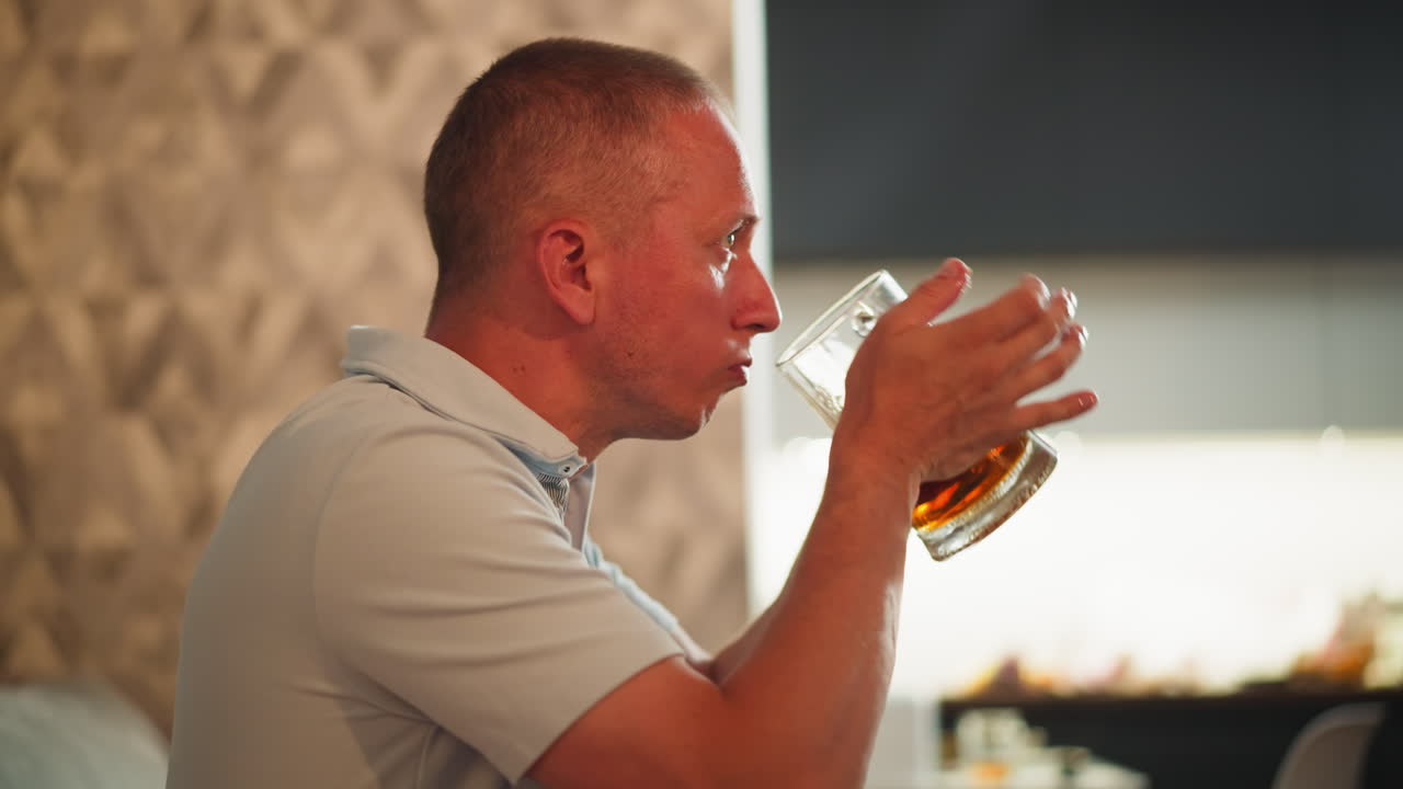 Man eating meal lifts glass of juice to drink and drops it back with satisfied look while sitting indoors, textured wall background, relaxed mood, enjoying refreshing beverage after food