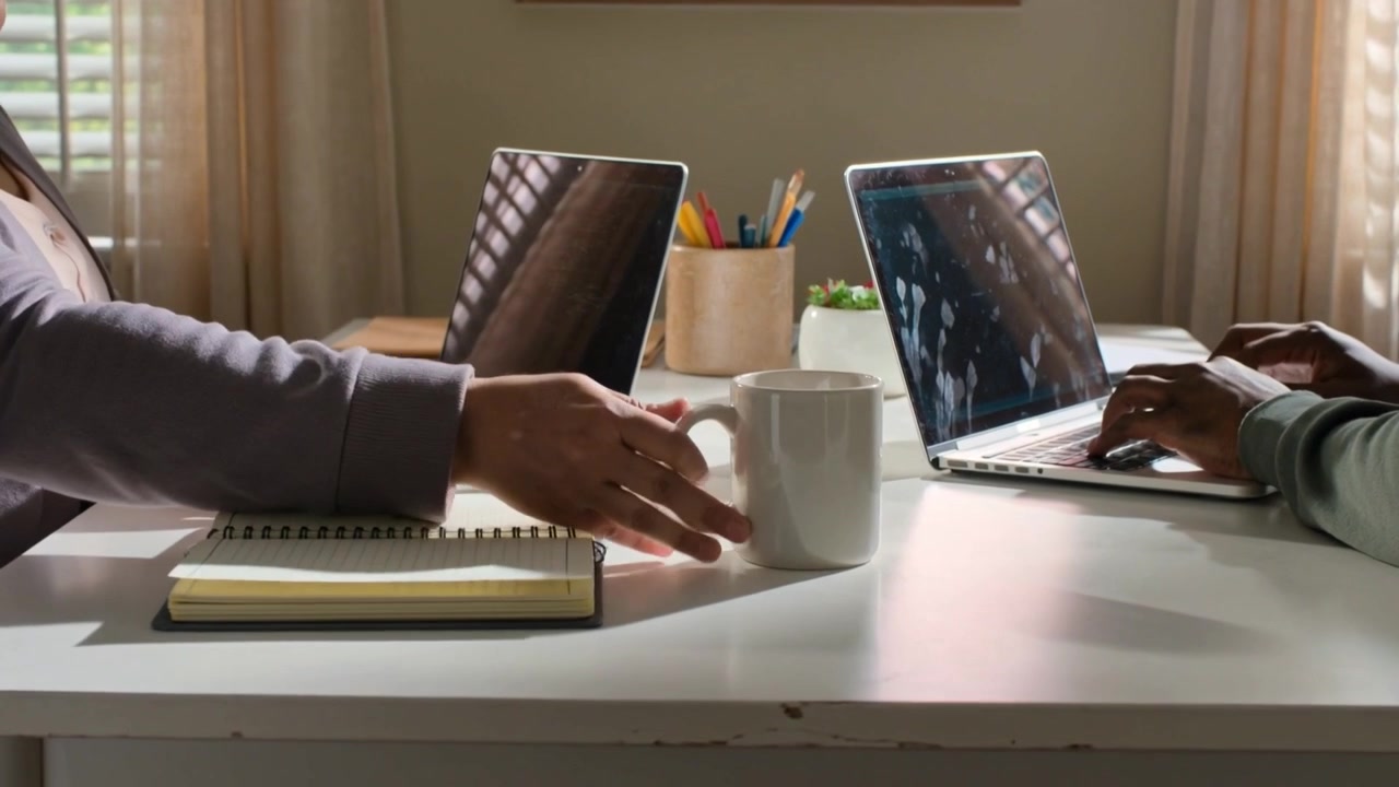 Flipping notebook, left grabbing mug to sip while right typing on laptops at home table