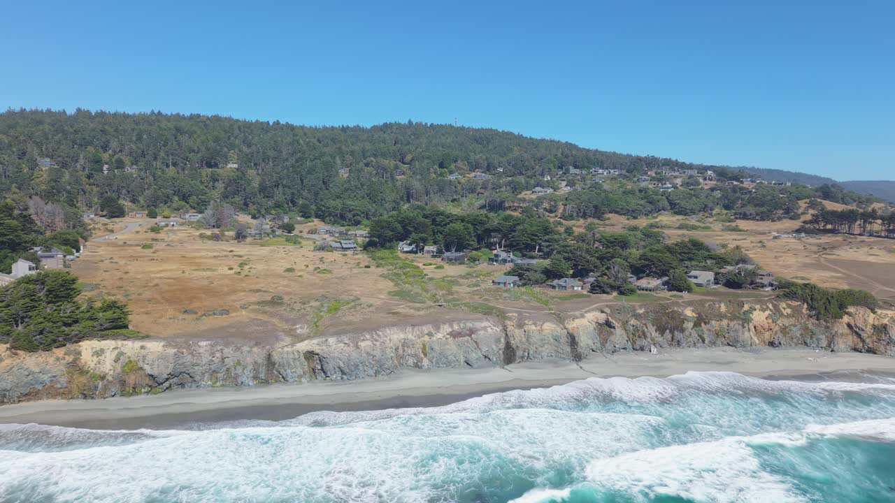 Drone panning shot from the beach reveals cliffside homes, forested hills, and powerful waves breaking along the shore at Black Point Beach, California