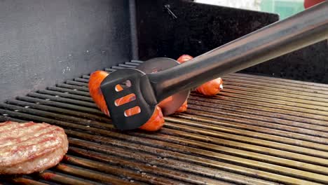 flipping gourmet hotdog weiner on hot bbq grill with steel tongs, close up slomo