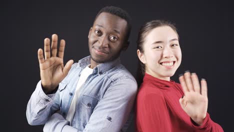 african man and asian woman looking at camera and waving in happy relationship.