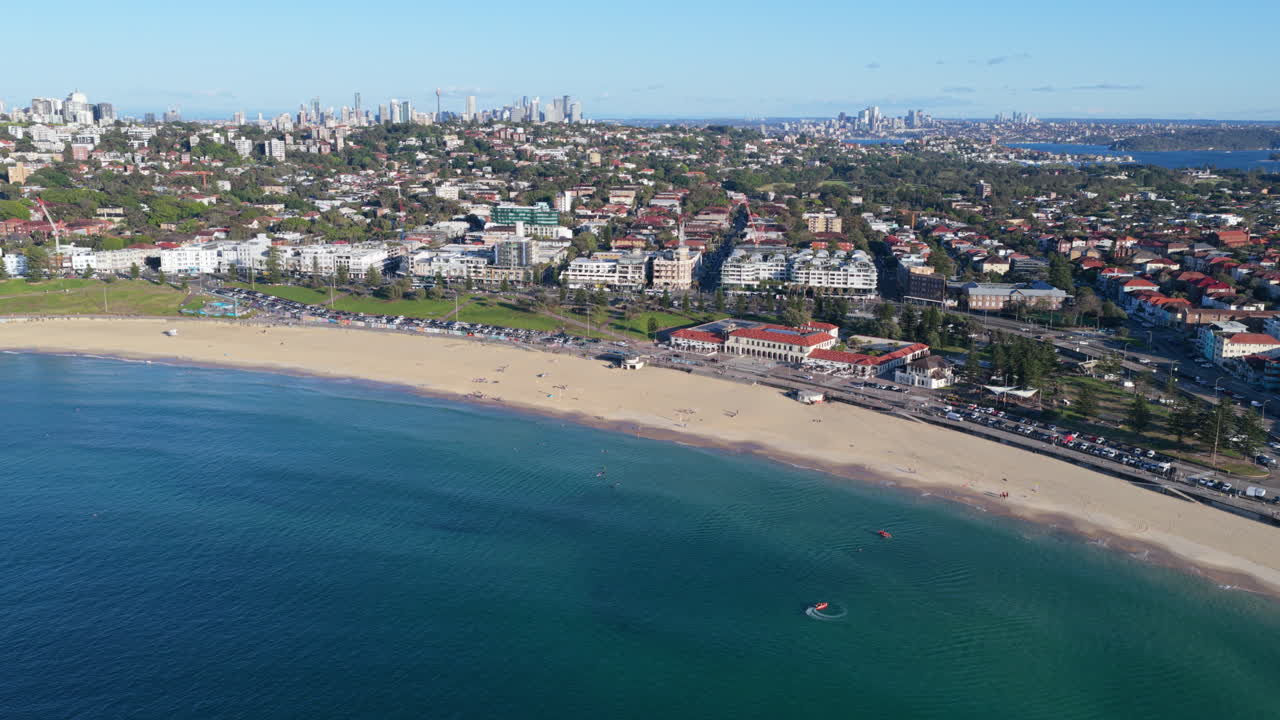 Eine weitläufige Luftaufnahme von Bondi Beach an einem sonnigen Sommermorgen mit ein paar Leuten, die surfen und sich am Strand aufhalten, sowie der weiten Skyline von Sydney und der städtischen Bebauung im Hintergrund