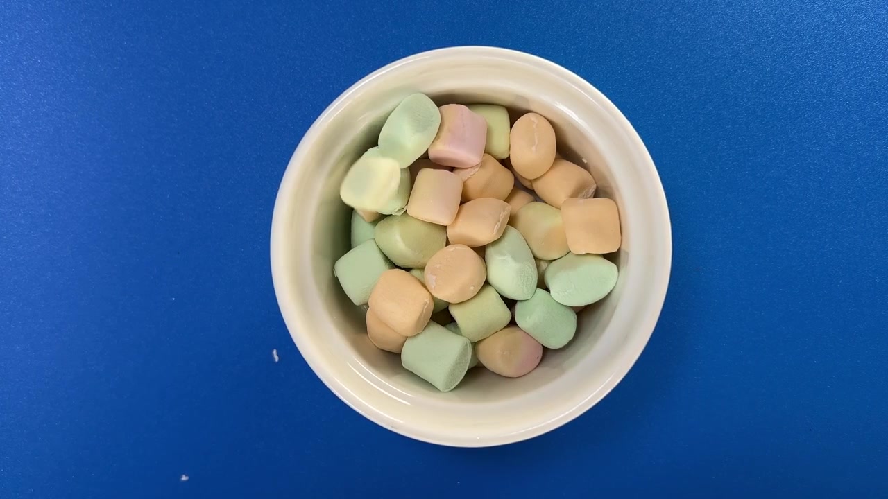 Marshmallow colorati che cadono in una ciotola bianca su sfondo blu, catturati a mezz'aria durante un'azione giocosa con pezzi multipli dai colori pastello.