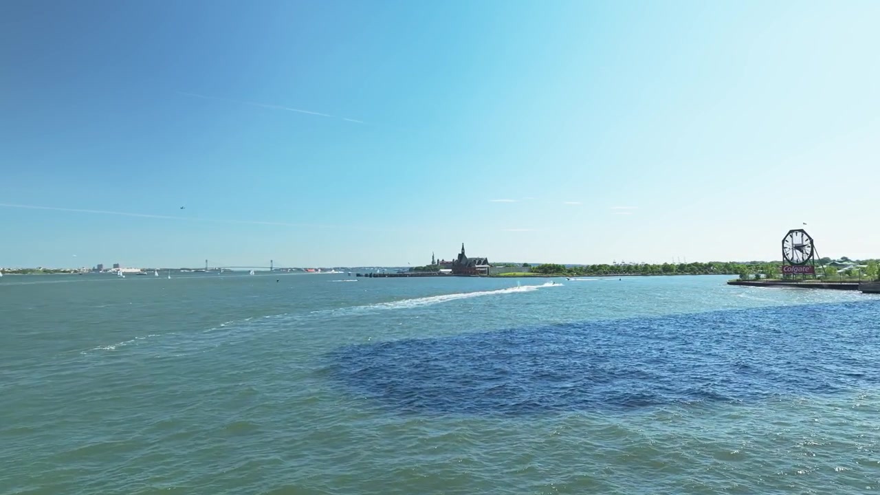Carrellata aerea verso Ellis Island con la Statua della Libertà in lontananza all'orizzonte sotto un cielo blu.