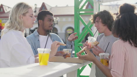 group of  friends sharing a pizza and drinking cold drinks standing at an outdoor table in the street, while chatting together 1