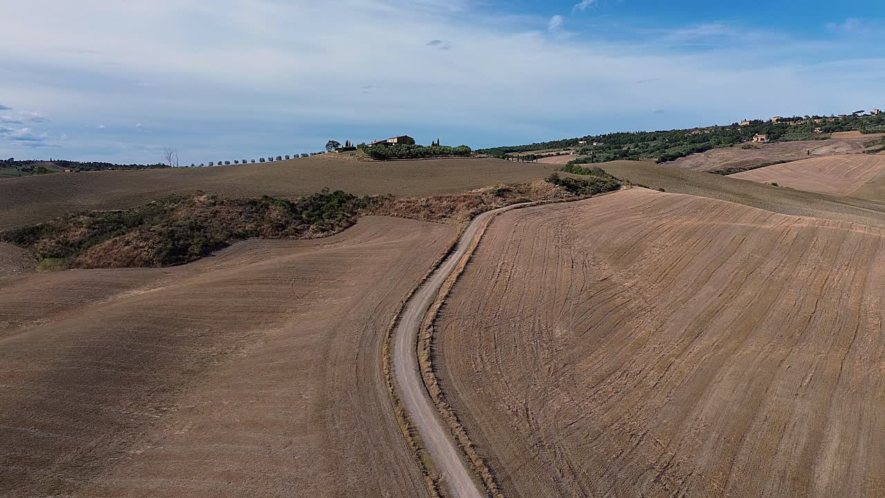 Aerial view of a winding dirt road cutting through harvested Tuscan hills with a distant cypress-lined estate under a blue sky.