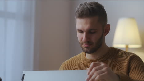 Un-Joven-Está-Abriendo-Una-Computadora-Portátil-Y-Comenzando-A-Trabajar-En-Casa-Como-Autónomo-Y-Trabajo-Remoto-Retrato-De-Primer-Plano-De-Un-Hombre-Caucásico-En-La-Mesa-De-La-Habitación