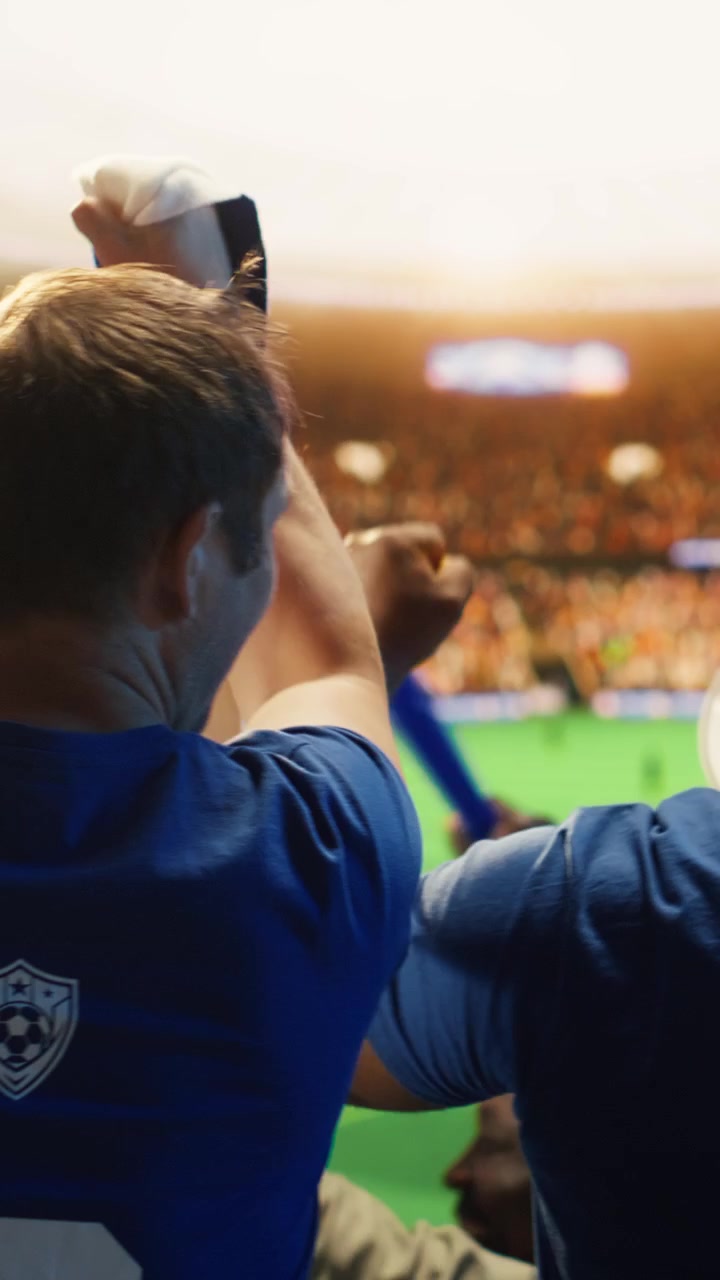 Video vertical. Aficionados al fútbol gritan con los brazos en alto en un estadio deportivo.