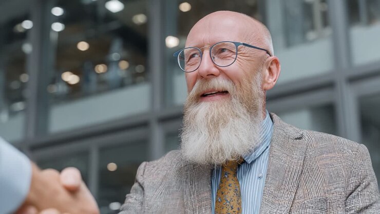 En glad forretningsmand med et distinkt skæg smiler, mens han giver hånd, symboliserende et succesfuldt partnerskab og gensidig aftale i en professionel ramme.