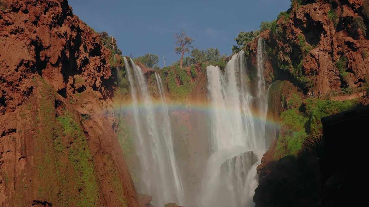Pelangi melengkung melalui semburan air di Air Terjun Ouzoud yang jatuh ke ngarai batu merah di Maroko