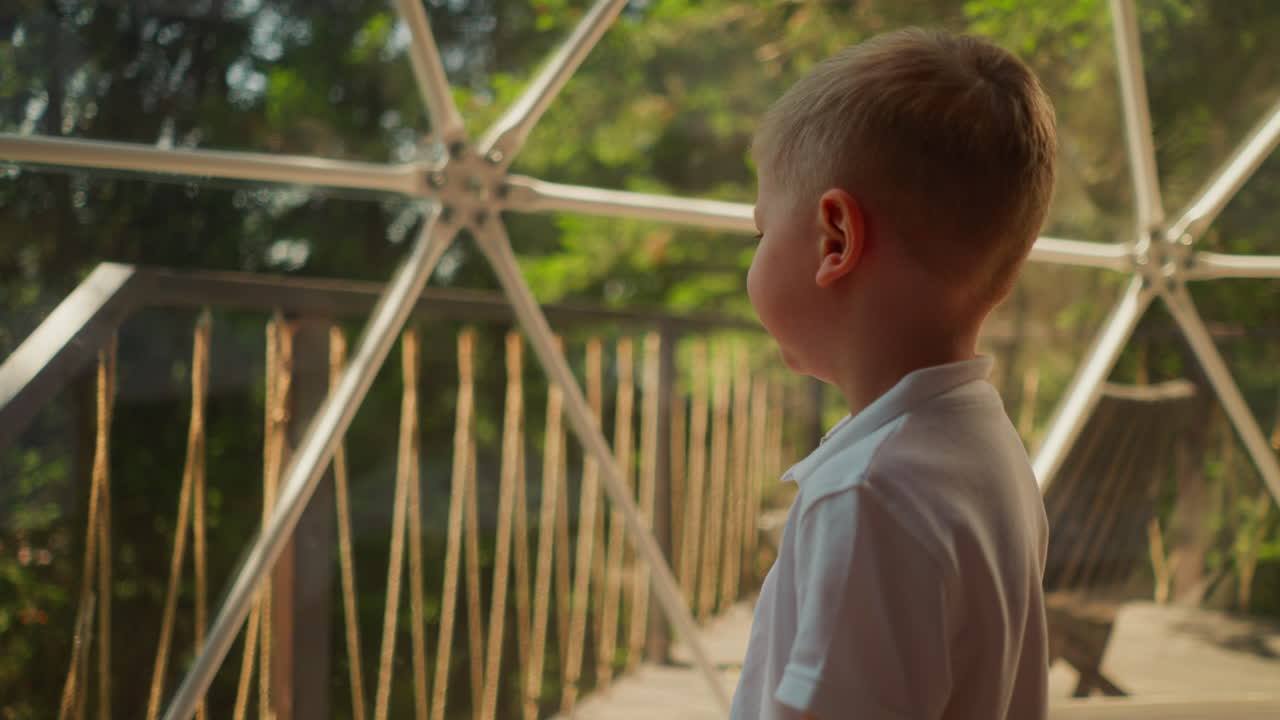 아이는 글램핑에서 프레임과 함께 유리 벽 근처에 서 있습니다. 작은 소년은  캐빈과 함께 델럭스 캠핑에서 휴식을 취합니다. 아이는 창문 근처의 산책로를 기다립니다.