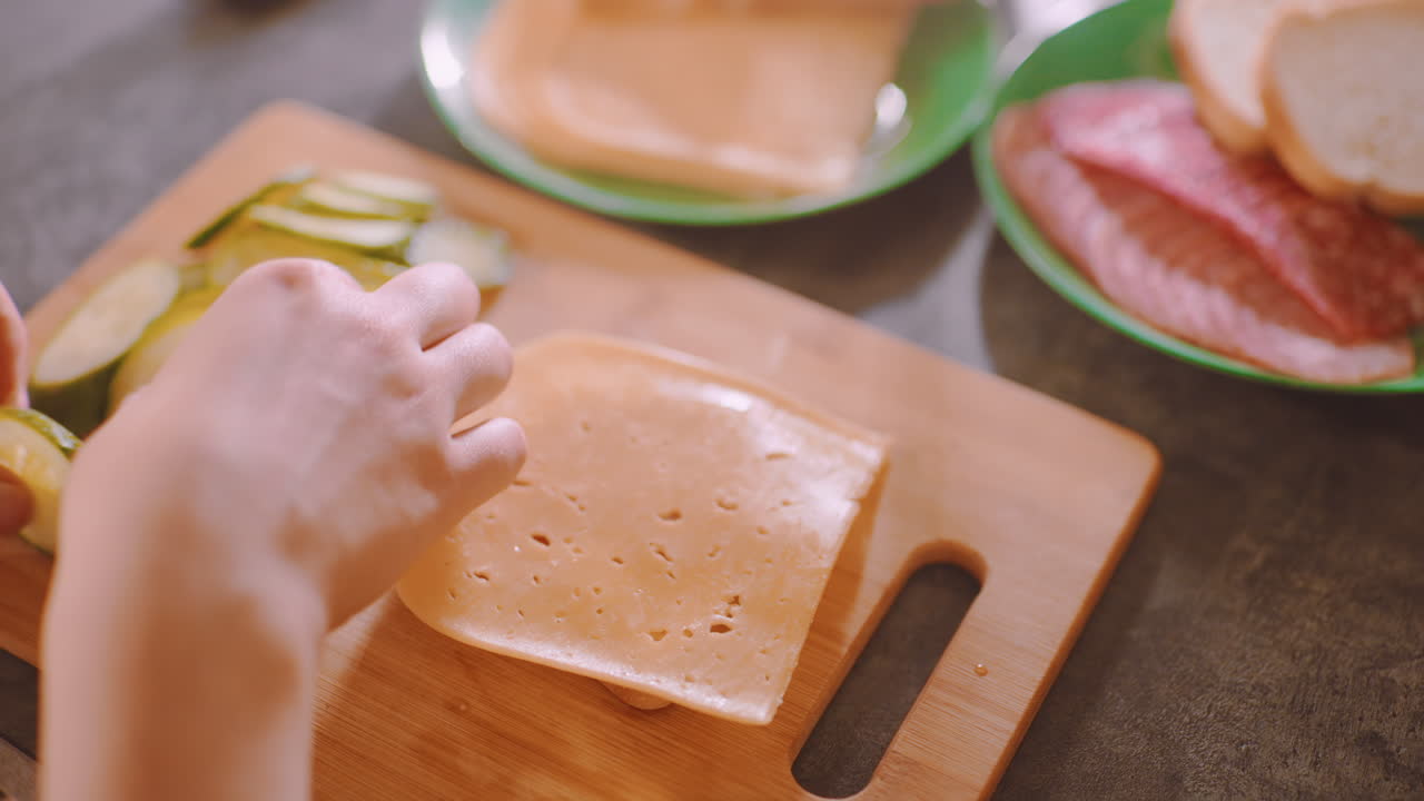 Close up of cook preparing breakfast with fresh bread, cucumber slices, cheese, and sausage on cutting board, arranging ingredients carefully for child meal beside cup of coffee