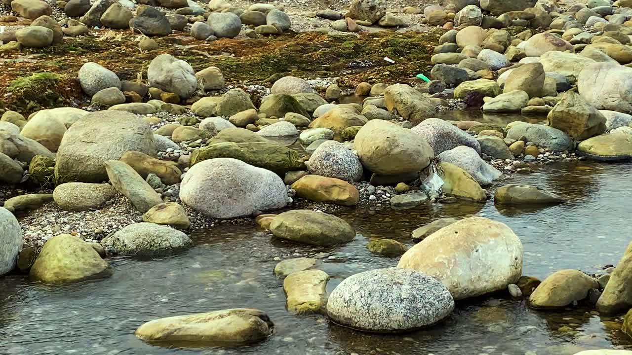 Sacchetto di politene incastrato tra le pietre del fiume in un ruscello poco profondo a Bichnakandi, Bangladesh. Primo piano sui rifiuti su ciottoli e massi che evidenziano l'inquinamento ambientale.