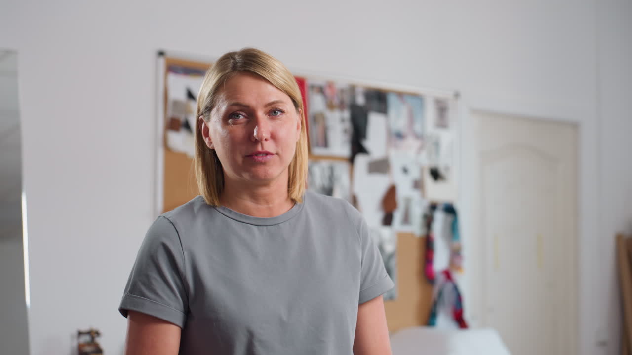 Playful needlewoman lifting black scissors pretending to cut material while smiling joyfully, standing in bright sewing workspace with creative boards and colorful textiles in background