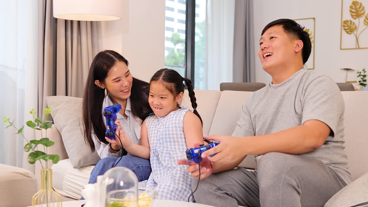 Parents and daughter laugh and play video games on couch, natural daylight, cozy home setting