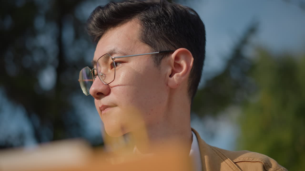 young man sketching outdoors with easel focused portrait study in sunny park, closeup facial shots, soft bokeh background, warm tones, deliberate pencil and brush strokes, urban trees in background