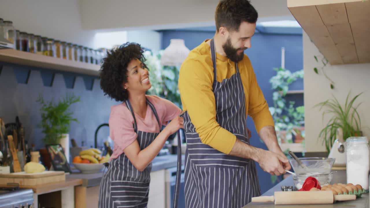 video de una feliz pareja diversa poniéndose delantales y preparando comida en la cocina en casa