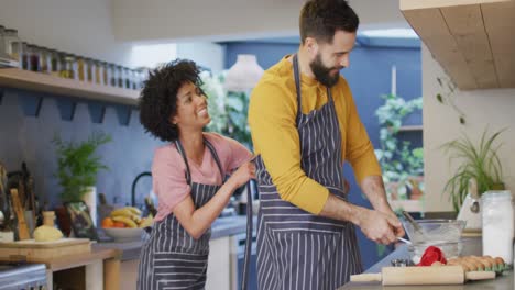 Video-De-Una-Feliz-Pareja-Diversa-Poniéndose-Delantales-Y-Preparando-Comida-En-La-Cocina-De-Casa