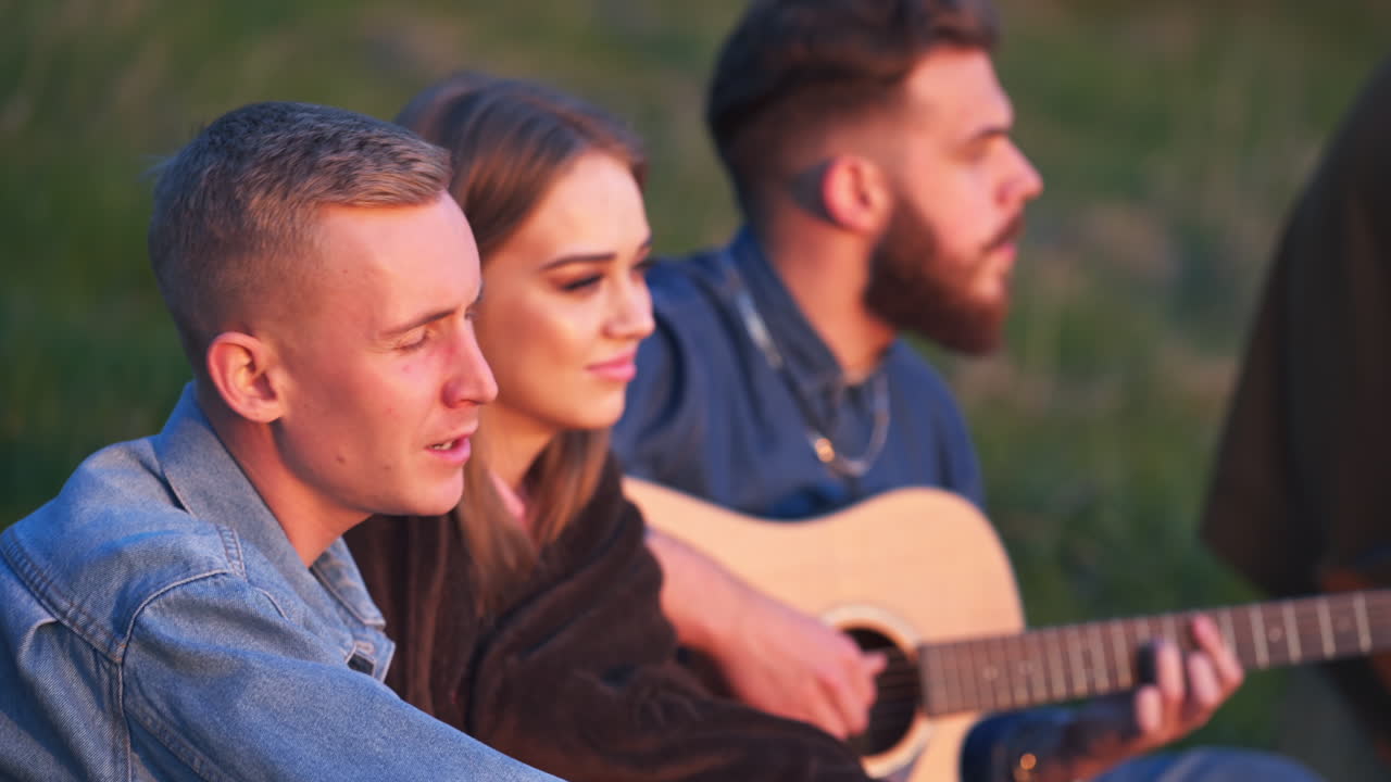 Friend sitting in the nature, singing songs under guitar music. Young people having rest and fun, their faces lit by the setting sun. Blurred backdrop.