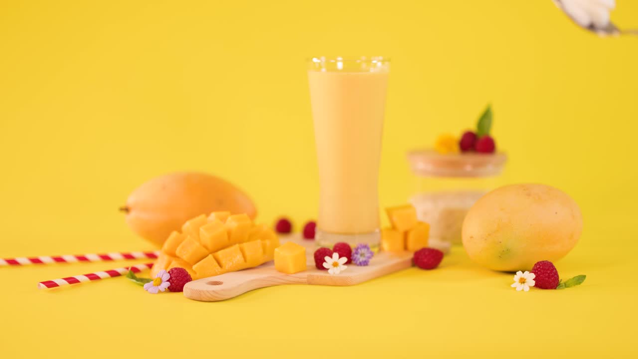 Una mano mette della panna montata vicino a un frullato tropicale al mango con frutta fresca su uno sfondo giallo brillante