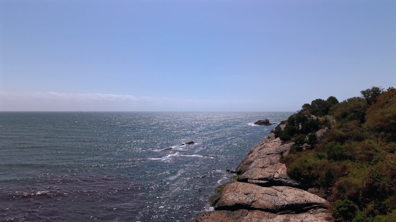 An aerial view of red tide, a reddish colored seaweed on the shore at Surfer's End in Middletown, RI on a sunny day with blue skies. The camera boom up by the rocks and green bushed by the shore