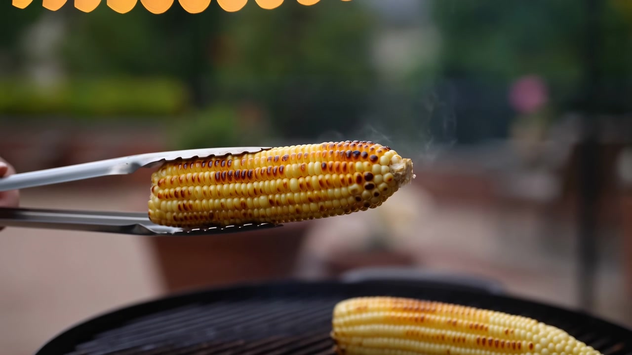 Close-up shows grilled corn cob held by serrated metal tongs above round barbecue grate while second cob rests below, golden kernels shining with charred spots, smoke, heat, summer cookout atmosphere