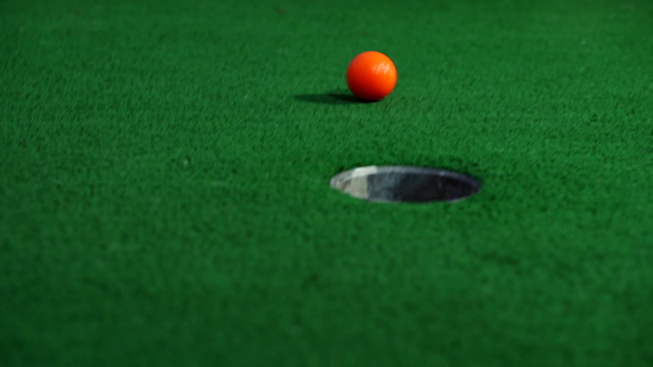 Una pallina da minigolf arancione esce da un tubo in un muro e rotola mancando la buca