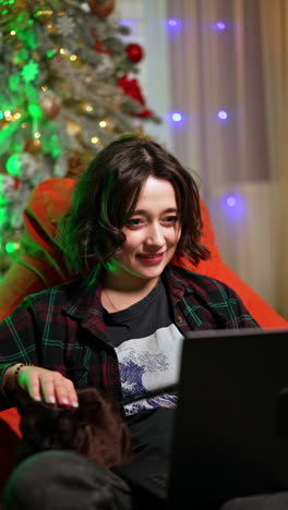 happy caucasian brunette sitting comfortably in the armchair pets her cat. girl is watching a film at home for christmas. vertical video.