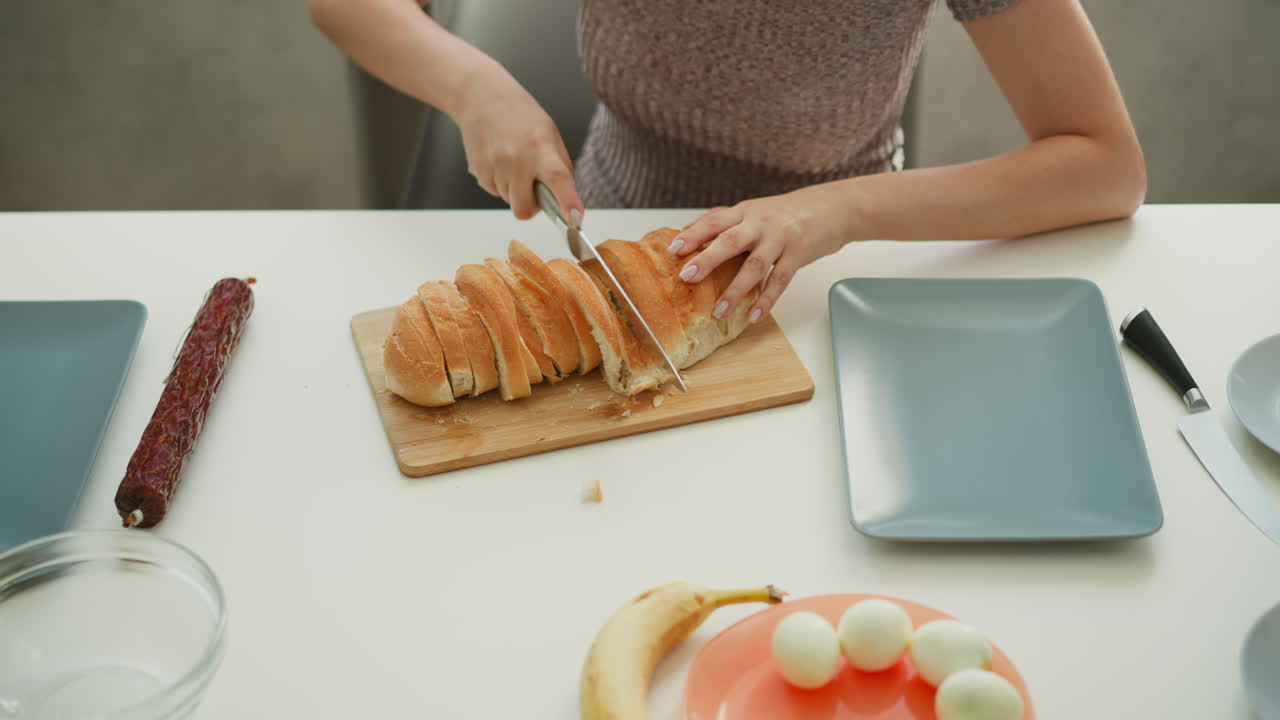 High angle hand view of female pastry cook cutting loaf of bread with knife on white table next to egg banana and bowls in kitchen under bright sunny morning light streaming through window behind