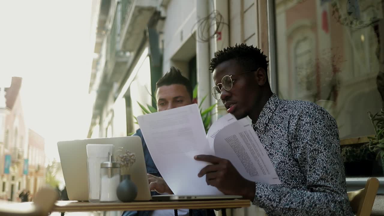 Multiethnic men working with papers and laptop in outdoor cafe