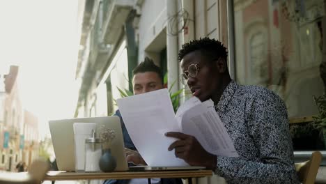 multiethnic men working with papers and laptop in outdoor cafe