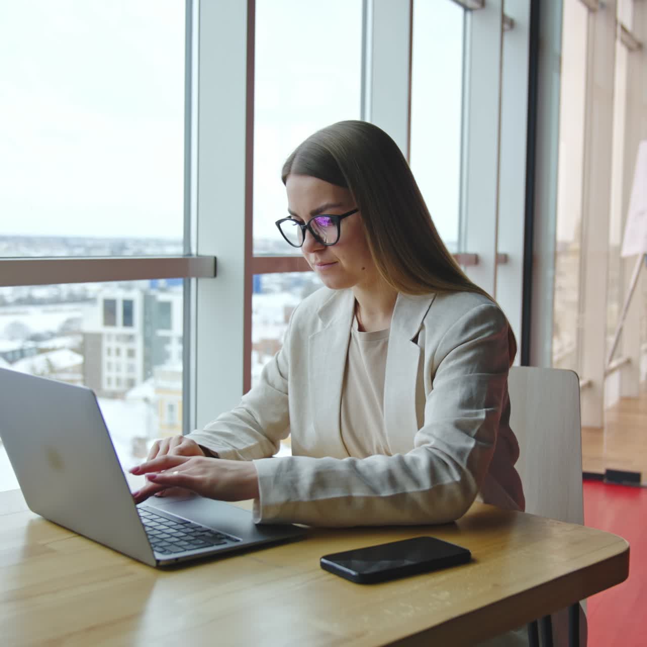 Smiling business lady sitting at the desk and working at laptop. Attractive woman at light office workplace. Panoramic windows with cityscape at backdrop