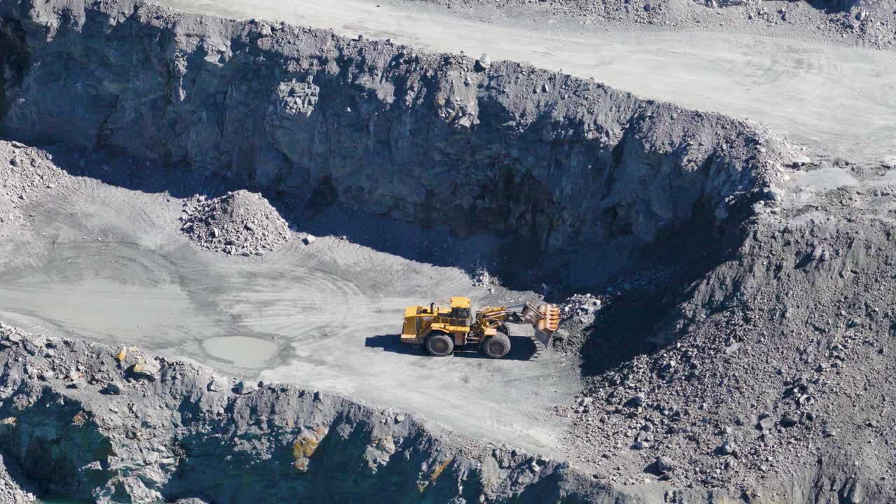 Telephoto drone captures excavator and trucks in action at a quarry. Bright daylight highlights the rugged terrain and industrial activity