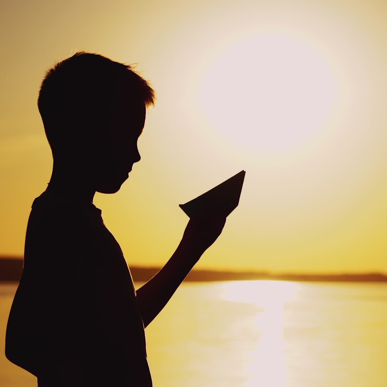 Little boy imitating swimming with his homemade boat near the river at sunset. Silhouette of a child with origami ship made from paper