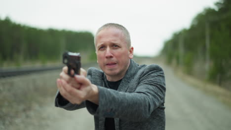 a close view of a man in a gray jacket aiming a handgun with a determined expression, the background shows a blurred railway track and forest