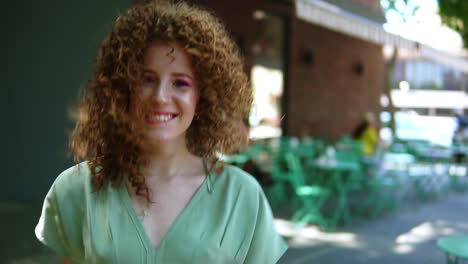 Beautiful-ginger-girl-with-perfect-hairdo---curls-walking-outdoors,-smiling-and-posing-to-the-camera-with-outdoors-cafe-terrace-on-background
