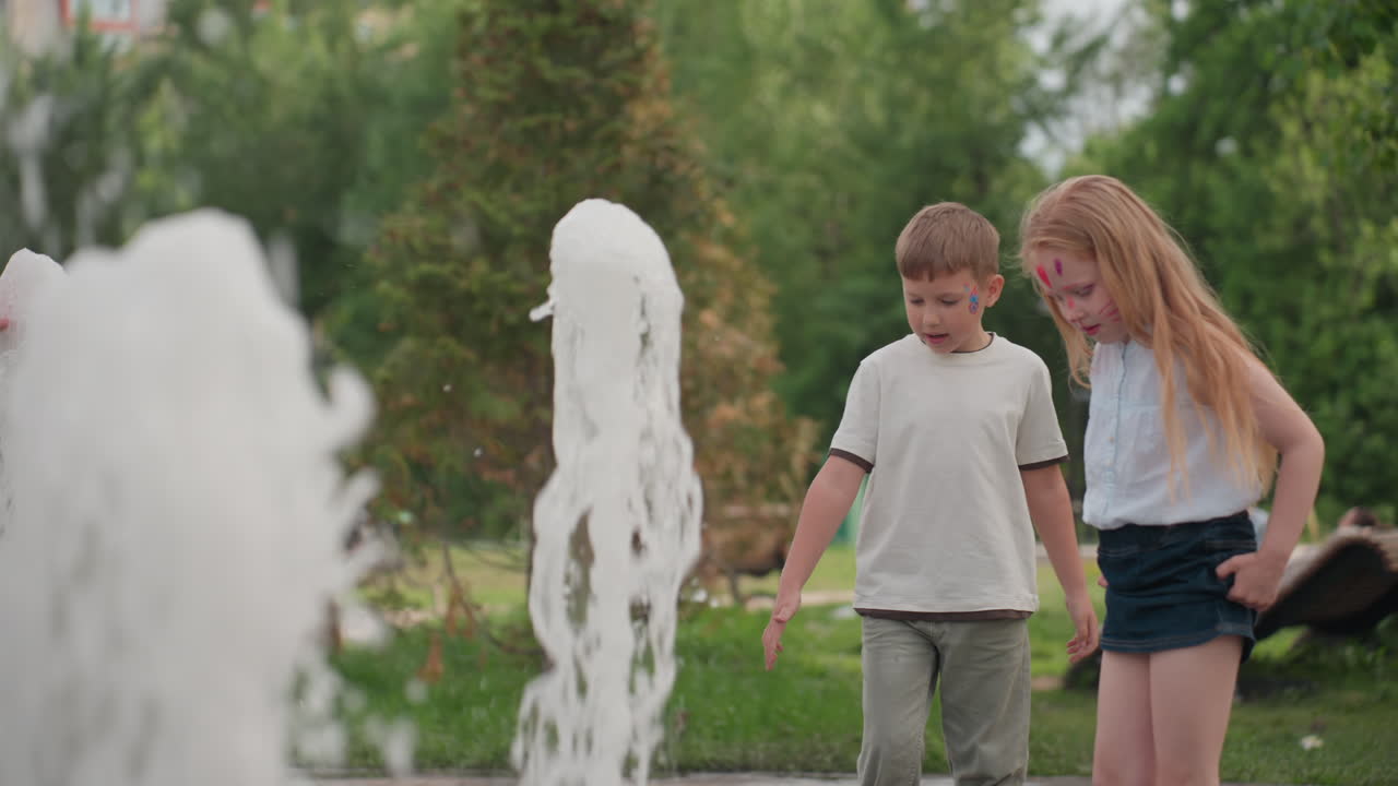 Toddlers playing with water jet in recreational park, touching splash with hands, parents and other people seated on bench in background, summer greenery framing curious youthful interaction