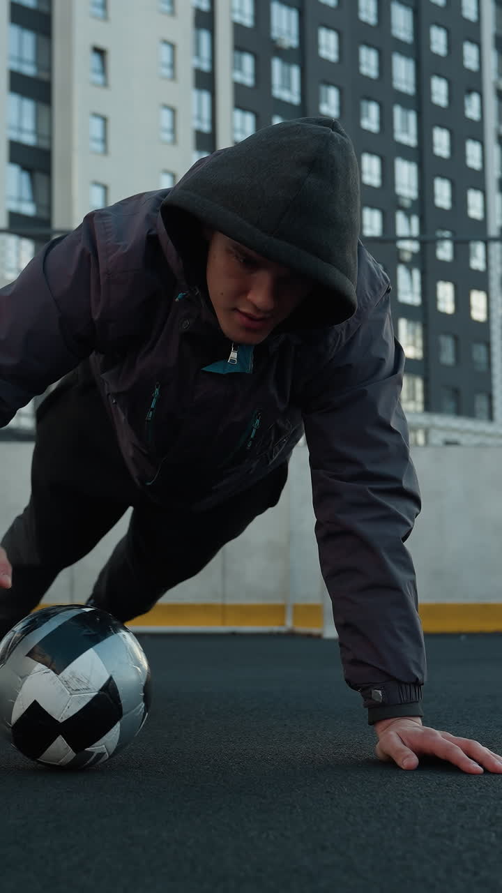 Young man performing dynamic push-ups alternating hand placement on soccer ball, showcasing strength, coordination, and agility, background includes goal post and residential buildings