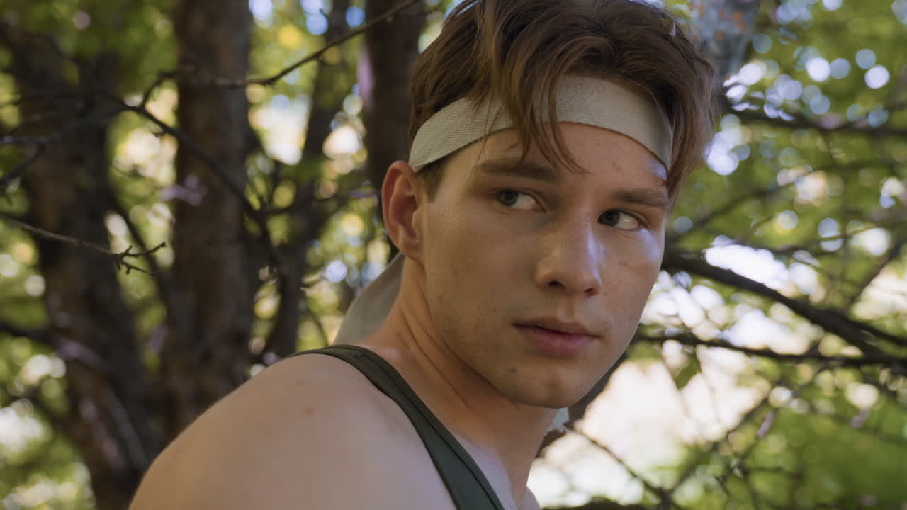 military soldier scanning forest canopy in alert side profile, headband tied, eyes rotating towards distant sound, tank top and visible tattoos, dappled light shadows across face, poised stance