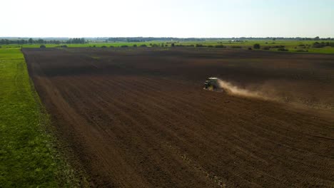 drone shot of a green john deere tractor preparing the field for plantation with a cloud of dust behind tractor on a sunny summer day, zooming in