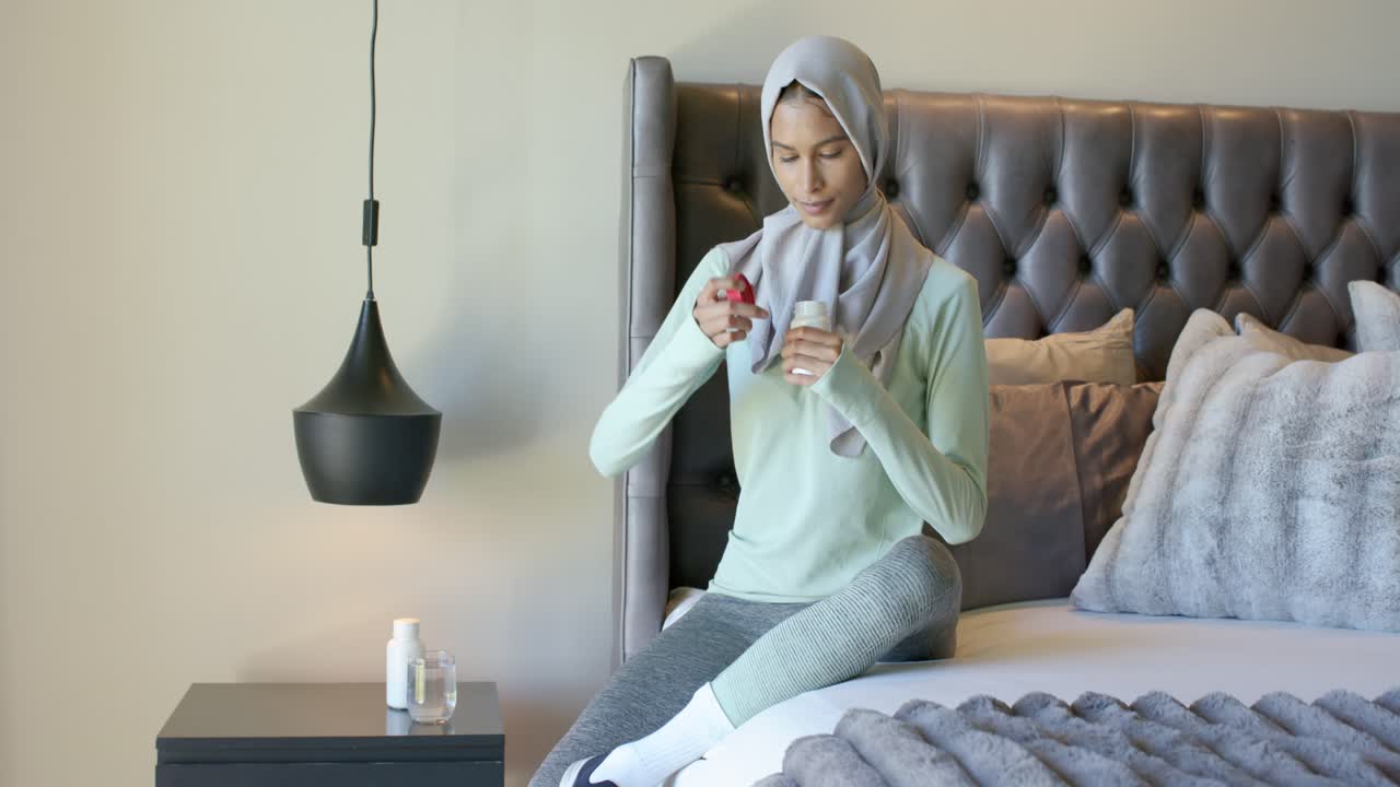 Donna afroamericana con hijab seduta sul letto che prende una bottiglia e applica una lozione per la cura delle mani.