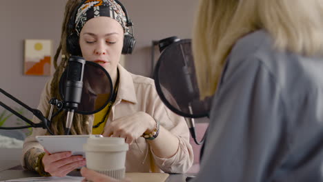 woman with dreadlocks recording a podcast talking into a microphone with another woman sitting at desk