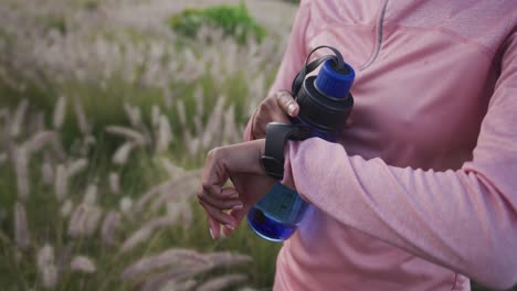 Midsection-of-mixed-race-woman-exercising-checking-smartwatch-during-a-run-in-countryside